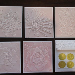 May include: Six embossed cards with floral designs. The cards are pink and white and feature a variety of flowers, including roses, daisies, and lilies.