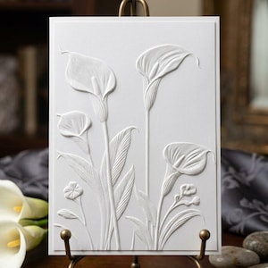 May include: A rectangular white card featuring a raised, embossed design of three calla lily flowers with long stems and pointed leaves. The card is displayed on a metallic bronze-toned easel stand, showcasing the intricate texture of the floral relief.