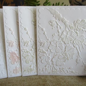 Puede incluir: Un juego de cuatro tarjetas de felicitación blancas con borde festoneado. Las tarjetas presentan un diseño floral en relieve con delicados acentos rosas y verdes. El diseño incluye flores y ramas.