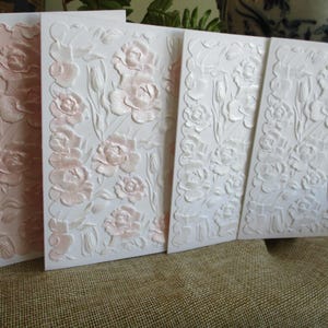 Puede incluir: Cuatro tarjetas de felicitación blancas con diseños florales en relieve. Las tarjetas presentan un patrón repetido de rosas y hojas, con un sutil tono rosa en las flores. Las tarjetas están dispuestas de forma ligeramente superpuesta.