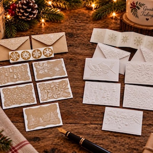 May include: A collection of rectangular greeting cards and envelopes featuring embossed winter designs. The set includes tan cards with white snowflake and forest patterns, and white cards with raised botanical and snowy landscape motifs.