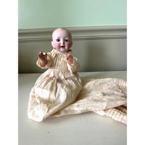 May include: A vintage porcelain baby doll with a serene expression, dressed in a cream-colored, pleated gown. The doll's arms are slightly raised, and it is resting on a matching fabric. The doll has rosy cheeks and a bald head.