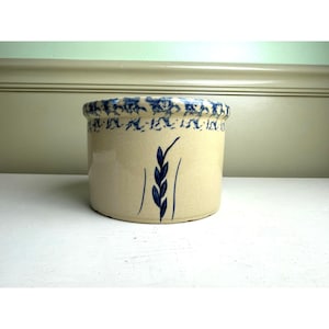 May include: A cylindrical ceramic crock with a beige base color, featuring a blue hand-painted wheat stalk motif on the front and a decorative blue floral pattern encircling the top rim. The vessel has a smooth, glazed finish throughout.