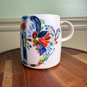 May include: White ceramic mug with a colorful floral design. The mug features a curved handle and a cylindrical shape. The floral pattern includes red, blue, and yellow accents. The mug is sitting on a wooden surface.
