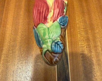 May include: A ceramic wall pocket in the shape of a stylized flower. The flower has red petals, a yellow center, and green leaves with blue accents. The base is brown. The wall pocket is mounted on a wooden surface.