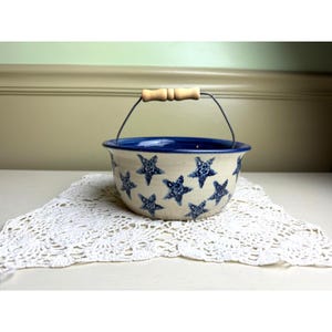 May include: A ceramic bowl with a cream-colored base featuring a pattern of blue speckled stars. The bowl has a dark blue interior rim and a metal wire handle with a light-colored wooden grip centered at the top for carrying the vessel.