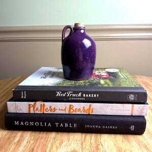 May include: A small, deep purple ceramic jug with a handle and cork stopper, resting on a stack of cookbooks. The cookbooks include titles like "Red Truck Bakery Cookbook," "Platters and Boards," and "Magnolia Table."