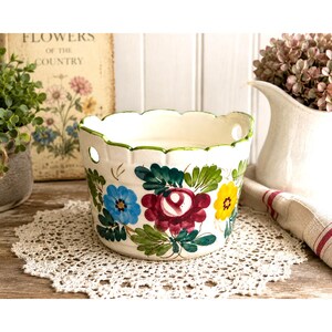 May include: A white ceramic bowl with a scalloped rim and two side holes, featuring hand-painted floral motifs in blue, deep red, and yellow with green leaves. The bowl has a glossy finish and sits on a white crocheted lace doily.
