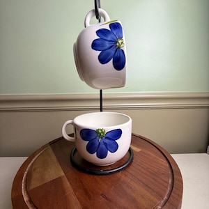 May include: Two white ceramic coffee mugs with blue floral designs. One mug is suspended upside down, while the other rests on a wooden surface. The mugs are held by a black metal stand. The wooden surface is round and has a dark brown color.