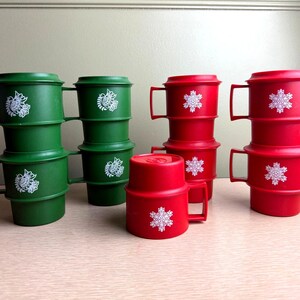 May include: Green and red plastic mugs with handles and lids. Each mug features a white snowflake design. The mugs are stacked in groups, suggesting a set for holiday use.