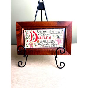 May include: A framed artwork with a dark wood frame, featuring the word "Dance" in red script. The artwork includes musical notes, a ballerina, and the quote "Dance is the Beauty, the inspiration, the energy and the joy of Music in motion."