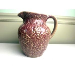 May include: A ceramic pitcher with a glossy mauve finish, featuring a raised, embossed relief pattern of grape clusters and vine leaves covering the entire exterior surface. The vessel includes a curved handle and a molded pouring spout.
