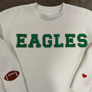 Könnte beinhalten: Ein weißer Langarm-Sweatshirt mit dem Wort "EAGLES" in grünen Buchstaben mit goldenen Verzierungen. Der Sweatshirt hat einen Fußball auf einem Ärmel und ein rotes Herz auf dem anderen.
