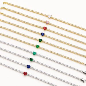 May include: A collection of tennis bracelets featuring a single heart-shaped gemstone set in the center of a row of small round clear stones. The bracelets are available in gold and silver tones with various colored heart stones including red, blue, green, pink, and clear.