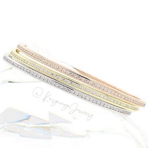 May include: Three gold bangles with a row of clear cubic zirconia stones on each. The bangles are stacked on top of each other, with the largest bangle on the bottom and the smallest bangle on top. The bangles are made of rose gold, yellow gold, and white gold.