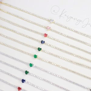 May include: A set of gold chain bracelets with a heart-shaped gemstone at the end of each chain. The gemstones are in various colors, including red, pink, green, and blue. The bracelets are all the same length and have a lobster clasp closure.
