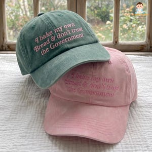 May include: A sage green baseball cap and a light pink baseball cap, both featuring the embroidered text 'I bake my own Bread & don't trust the Government' in a matching thread color. Each cap has a curved brim and a structured six-panel design.
