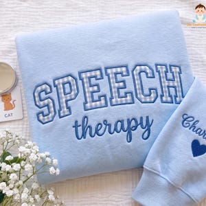 May include: A light blue sweatshirt featuring the embroidered text SPEECH in a blue and white gingham pattern, with the word therapy in blue script below. The sleeve is embroidered with the name Charlotte and a small blue heart icon.