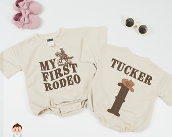 May include: Two beige baby bodysuits with short sleeves. One says "MY FIRST RODEO" with a cowboy graphic. The other says "TUCKER" with a cowboy hat graphic. Pink baby shoes and sunglasses are also in the image.