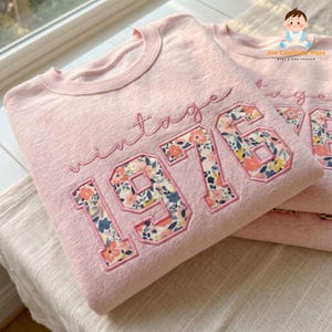 May include: A light pink crewneck sweatshirt featuring the word vintage in cursive script above the numbers 1976. The numbers are filled with a floral pattern of orange, yellow, and blue flowers on a white background with pink stitched outlines.