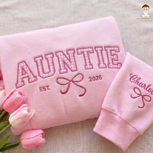 May include: A light pink sweatshirt featuring the embroidered text AUNTIE in a pink and white gingham pattern, with EST. 2026 and a bow icon. The sleeve displays the name Charlotte embroidered in pink above a small bow icon.
