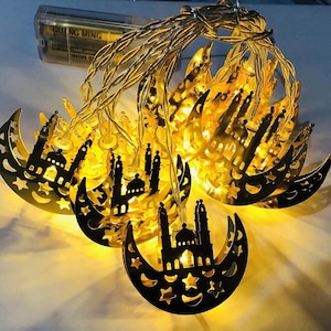 May include: A string of golden crescent moon-shaped fairy lights with a mosque silhouette design. The lights are strung together with clear wire and powered by batteries.