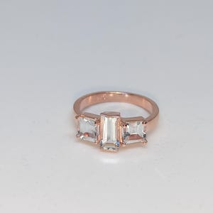 May include: Rose gold ring with three clear, rectangular gemstones. The central stone is vertical, with two horizontal stones either side. The ring is set against a white background, highlighting the design and sparkle of the jewellery.