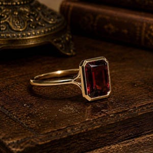 May include: A gold ring featuring a large, rectangular, deep red gemstone set in a smooth bezel setting. The band is thin and polished, connecting to the stone setting with a split shank design that highlights the vibrant, dark red stone.