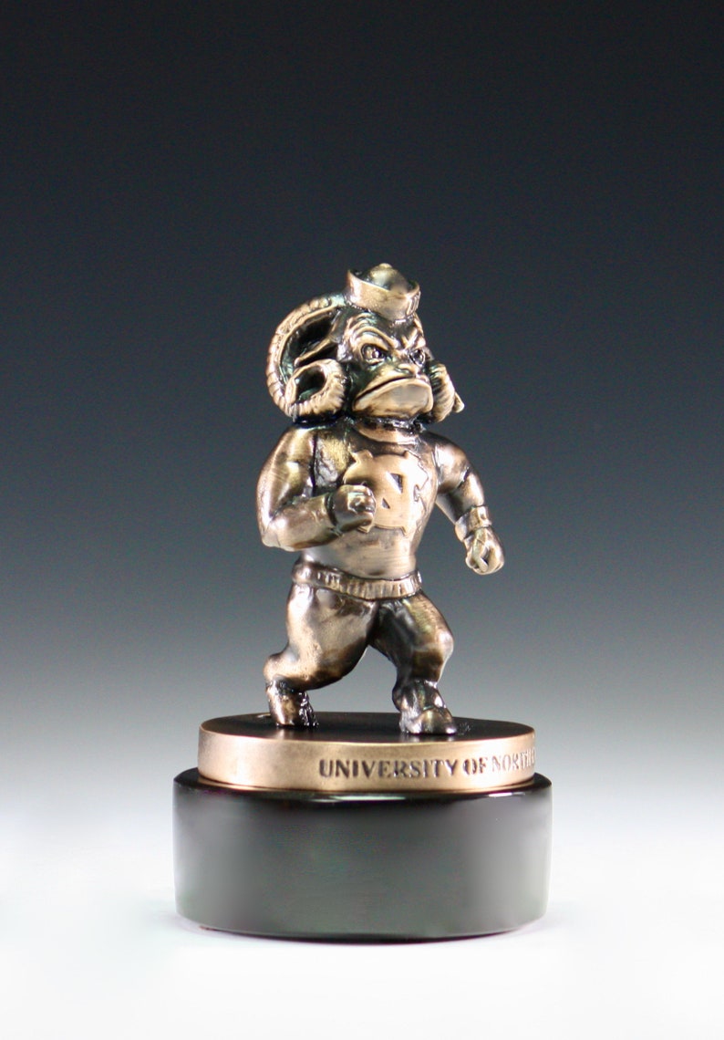 Puede incluir: Una estatua de bronce de una mascota de carnero con un sombrero de marinero y una camiseta con el texto "UNIVERSITY OF NORTH" en la base.