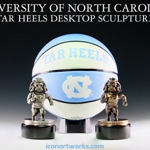 Puede incluir: Una escultura de escritorio de los Tar Heels de la Universidad de Carolina del Norte en plata y bronce con un bal&oacute;n de baloncesto y dos mascotas. El bal&oacute;n de baloncesto es azul y blanco con las palabras "Tar Heels" impresas en &eacute;l. Las mascotas est&aacute;n de pie sobre bases negras con las palabras "University of North Carolina" impresas en ellas.