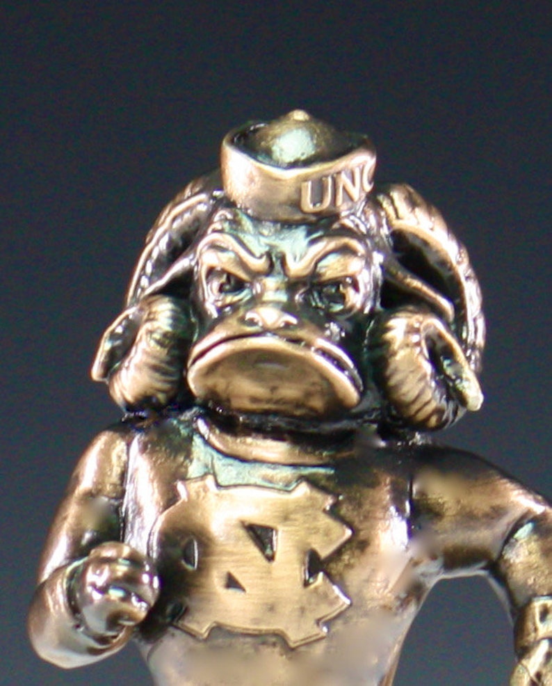 Puede incluir: Una figurita de bronce de una mascota que lleva un gorro de marinero con las letras "UNC" en &eacute;l. La mascota tiene una cara grande y expresiva y lleva una camisa con un gran logotipo de "UNC".