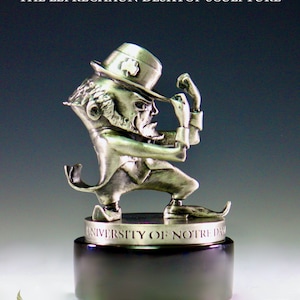 May include: A silver-colored desktop sculpture of the University of Notre Dame leprechaun mascot. The leprechaun is wearing a green hat with a shamrock and is holding his hat in his right hand. The sculpture is on a black base with the university's name inscribed on it.