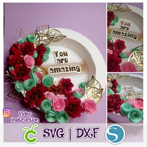 May include: A white circular frame with the words "You are amazing" in gold letters surrounded by a wreath of red, pink, and green paper flowers. The frame is on a purple background.