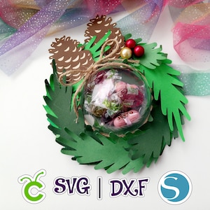 Op de afbeelding: Een groene papieren kerstkrans met dennennaalden en bruine dennenappels. Een doorzichtige plastic bol gevuld met kleurrijke snoepjes bevindt zich in het midden van de krans. De krans is vastgebonden met touw en versierd met rode bessen.