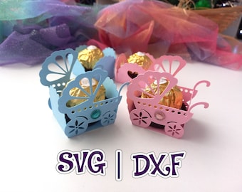 May include: Two paper baby carriage favor boxes, one blue and one pink, with a chocolate candy inside each. The boxes are decorated with cut-out designs and have a small rhinestone on each wheel. The text "SVG | DXF" is printed below the boxes.