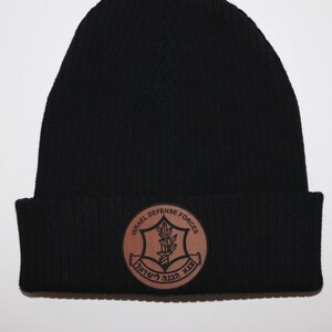 May include: Black knit beanie with a brown leather patch featuring the Israel Defense Forces logo. The logo is a Star of David with a sword and olive branches inside. The text "ISRAEL DEFENSE FORCES" is written around the star.