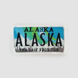 Puede incluir: Un parche bordado blanco, azul y verde con el texto "ALASKA" y "THE LAST FRONTIER" y un gráfico de una cordillera.