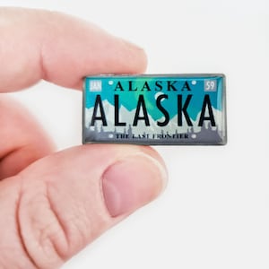 May include: A rectangular Alaska license plate magnet. The magnet features a teal and blue background with the word "ALASKA" in large white letters. The text "THE LAST FRONTIER" is at the bottom. The magnet is held by a person.