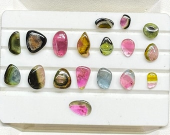 May include: A collection of small, polished, multi-colored tourmaline gemstones arranged on a white display tray. The stones feature various hues including pink, green, yellow, blue, and black, each with a smooth, rounded, and irregular shape.