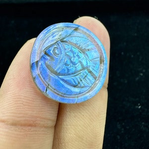 Op de afbeelding: Een ronde, blauwe labradorit-cabochon met een gegraveerd visontwerp. De edelsteen heeft iriserende blauwe en groene kleuren.