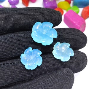 May include: Three light blue flower-shaped cabochons with a carved design. The cabochons are made of a translucent material and have a slightly iridescent sheen.