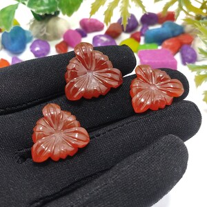 May include: Three carved, reddish-orange gemstone flower pendants. Each pendant features a detailed floral design with multiple petals and a central point. The pendants are displayed on a black surface, with colorful stones and greenery in the background.