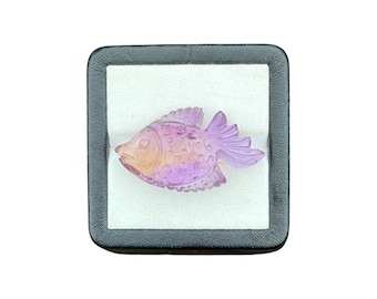 May include: A carved gemstone fish figurine featuring a gradient of translucent purple and soft orange hues. The fish has detailed etched scales and fins, presented inside a square black display box with a white interior lining for contrast.