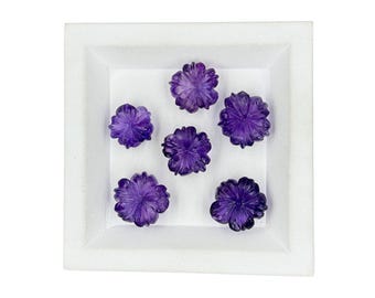 May include: Six carved purple amethyst gemstones shaped like five-petaled flowers. Each stone features intricate petal detailing and a polished finish. The gemstones are arranged in a cluster within a square white display tray with raised edges.