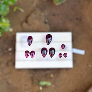 May include: An assortment of polished, teardrop-shaped gemstones in varying sizes. The gems are a deep, rich burgundy color, with a glossy surface. They are arranged on a white rectangular surface, set against a blurred background.