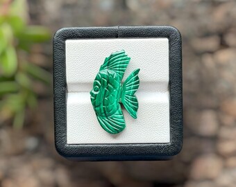 May include: A carved malachite gemstone shaped like a fish, featuring deep green and light green banded patterns. The fish carving displays detailed etched lines on its fins and body, showcasing the natural swirling texture of the stone.