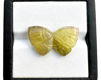 May include: Two yellow-green gemstone butterfly wings, carved with detailed wing patterns, are displayed in a black box with a white interior. The gemstones have a translucent quality, and the carving is intricate.