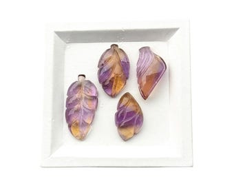May include: Four carved gemstone pendants in a leaf shape, featuring a gradient of purple and yellow hues. These translucent stones display intricate vein detailing on their surfaces, arranged together within a square white tray.