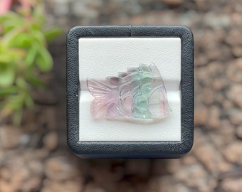 May include: A carved fluorite gemstone in the shape of a fish, featuring translucent bands of pale pink, soft green, and clear white. The fish carving is displayed on a white square insert within a black square frame, highlighting its details.