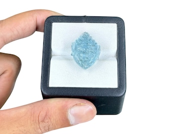 May include: A light blue, translucent gemstone carved into the shape of a face with detailed facial features. The gemstone is securely held within a white velvet insert inside a square, black display box with rounded corners and a smooth finish.